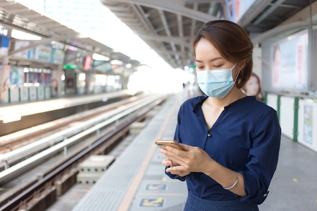 Asian woman in short hair uses smartphone while waiting train at station and wears medical face mask