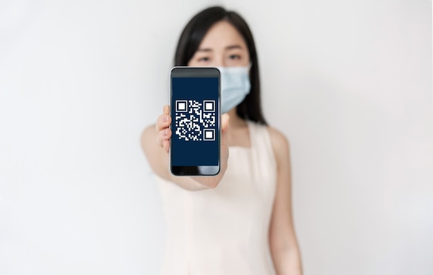 QR 코드 스캔 및 확인 기술을 화면에 표시하고 수술 용 얼굴 마스크를 착용 한 모바일 스마트 폰을 보여주는 아시아 여성