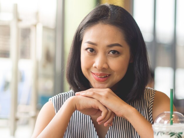 Asian woman sits in coffee cafe wirh iced coffee on table.