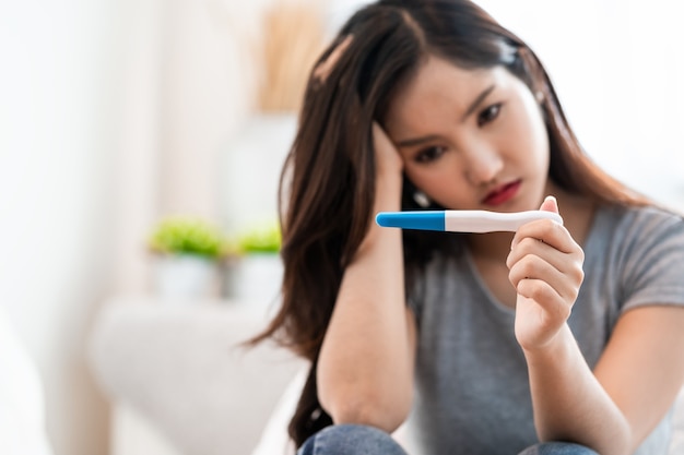 침대에 앉아 있는 아시아 여성은 임신 테스트 결과를 보고 실망감을 느낀다