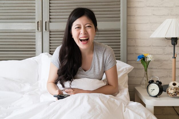 Asian woman sitting in the bed and watching the tv and burst\
into laughter while she see the funny movie.