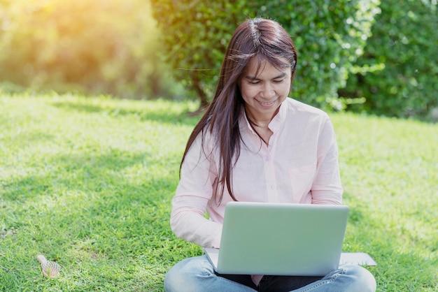 Asian woman sitting green park using laptop computer Woman working on laptop happy entrepreneur business using notebook with hands typing on keyboard home office during coronavirus quarantine period