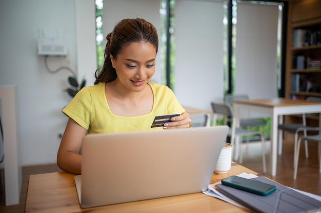 Foto donna asiatica seduta e con in mano una carta di credito per fare acquisti online con il portatile e lo smartphone a casa