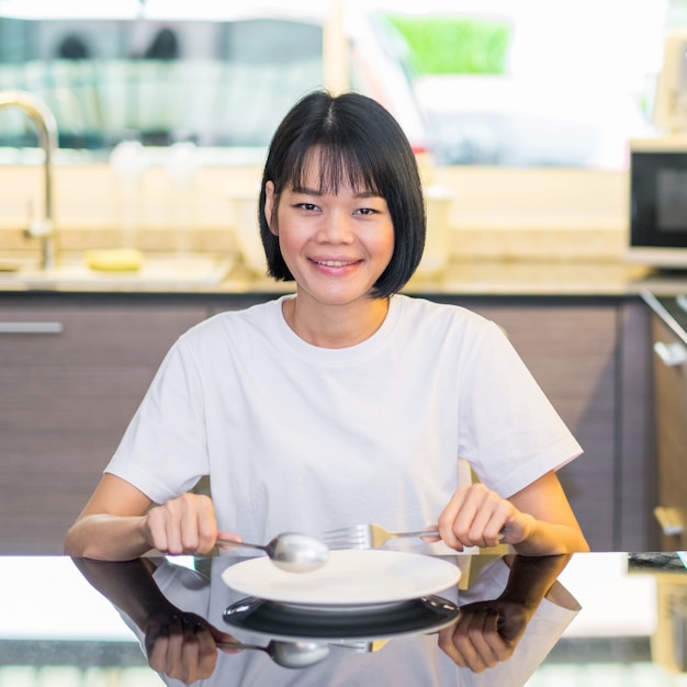 スプーンと白いお皿を持ってキッチンに座っているアジアの女性