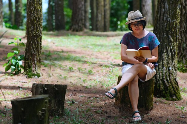 アジアの女性が公園で独りで丸太の上に座って読んでいる本で幸せに見えます