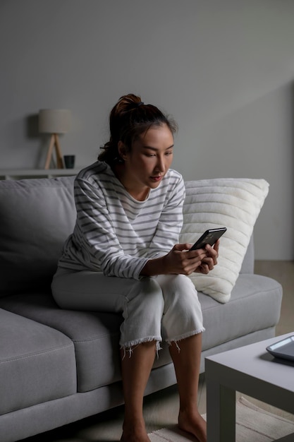 写真 ソファに座って電話を見ているアジア人女性 失望した気持ち 悲しい気持ち 女性 精神的な健康問題に苦しんでいます