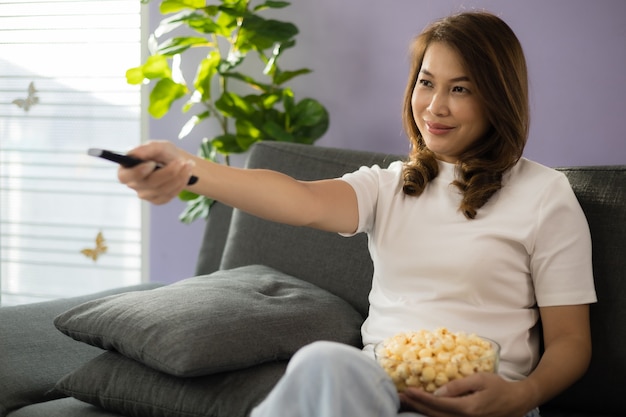 Азиатская женщина, сидящая на диване с расслабляющим и уютным жестом, держащая пульт дистанционного управления, нацеленная на телевизор, смотрит телевизор и улыбается весело и счастлива в гостиной с солнечным светом и легкой вспышкой.