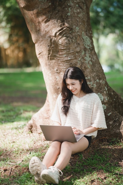 Asian woman sitting on picnic and lawn at park working on\
laptop asian female using laptop while sitting under a tree at park\
with bright sunlight work from anywhere concept