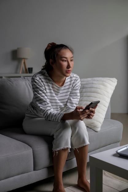 Photo asian woman sitting on sofa holding phone feeling disappointed sad upset female suffer from mental health problems