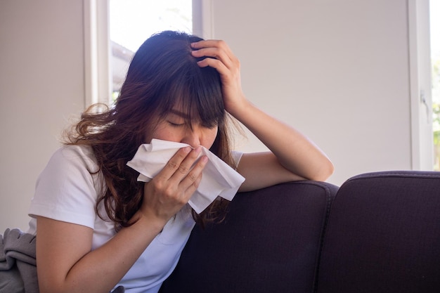 Asian woman sitting on the sofa inside the house having a\
fever, having a mouth cloth after covering the mouth after\
preventing spreading covid-19 is currently spreading around the\
world