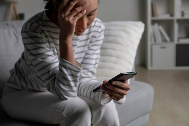 Foto donna asiatica seduta sul divano che guarda il telefono sentendosi delusa triste sconvolta femmina che soffre di problemi di salute mentale