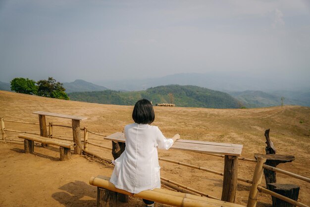 木製の椅子に座って山の景色を見ているアジア人女性の後ろに、風景を持つ人々。