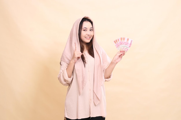 Asian woman smiles at the camera with her hand pointing up and carrying rupiah money for the competi
