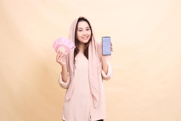 Asian woman smiles at you holding rupiah money and holding a cellphone gadget screen beautiful young