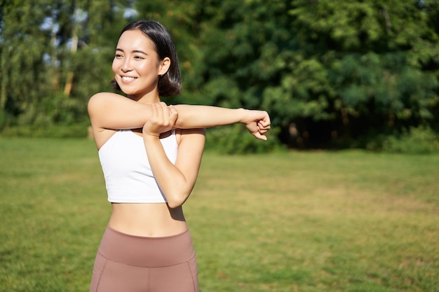 Asian woman stretching her arms doing fitness workout in park smiling pleased warming up before jogg