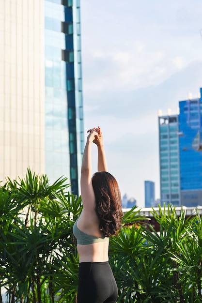 Asian woman strethes arms in active sport bra and leggings after work out in yoga pose relaxing her moment in rooftop city lifestyle everyday routine daily life with city view background