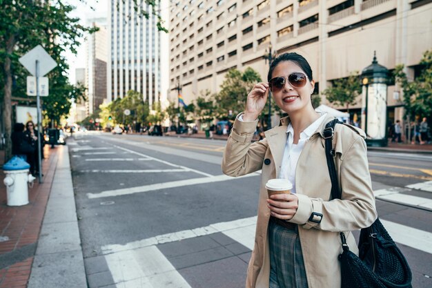 Photo asian woman in sunglasses smiling face camera. young elegant office lady drinking cocoa walking on zebra crossing in wide clean street with no car. young people city urban lifestyle in san francisco.