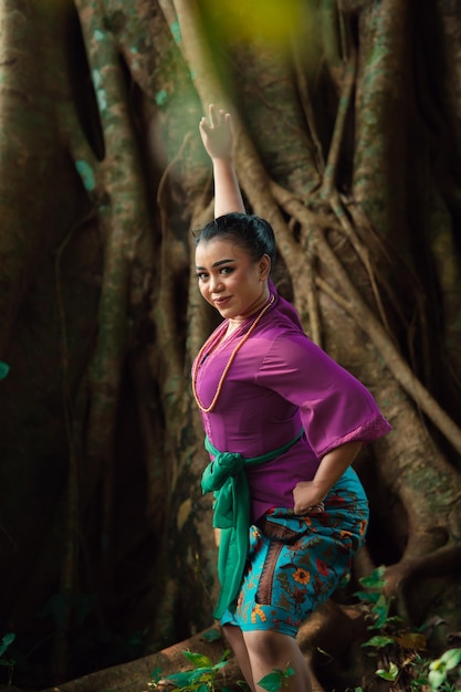 Asian woman takes a dancing pose in the jungle while wearing a purple dress and a golden necklace with a beautiful smile