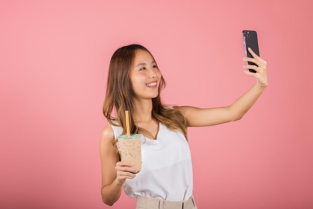 Asian woman takes photo of herself with drinking brown sugar flavored tapioca pearl bubble milk tea, smile female making selfie, studio isolated on pink background, pearl milk tea beverage concept