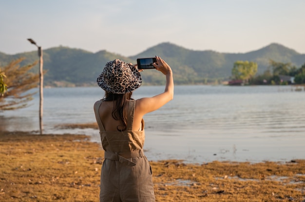 夕日の山の湖の景色でスマートフォンで写真を撮るアジアの女性