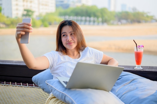 스마트폰으로 소셜 미디어를 위해 셀카 사진을 찍는 아시아 여성. 해변에서 온라인으로 일하는 사업가.