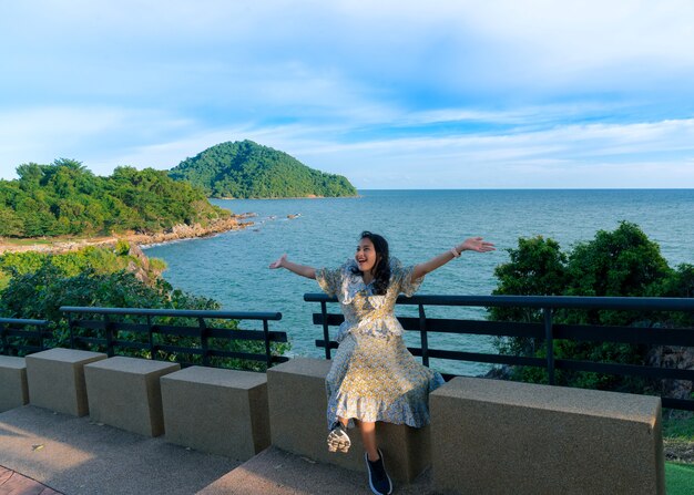 Asian woman tourist sit and feel fresh with happy arm raised at seat beside walkway on Noen Nang Phaya viewpoint with blue sea and mountain view, travel attraction at Chanthaburi, Thailand