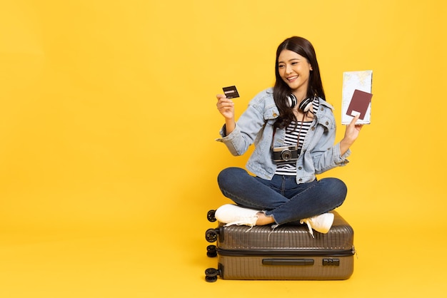Asian woman tourist sitting on baggage and holding credit card isolated on yellow background