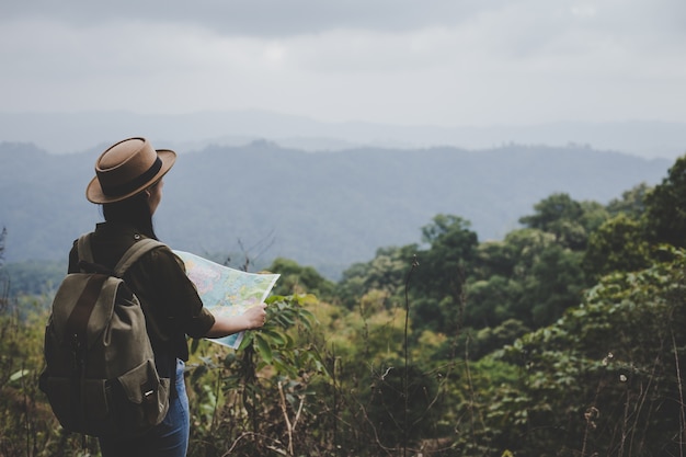 Asian woman traveler with backpack checks map to find directions in forest.