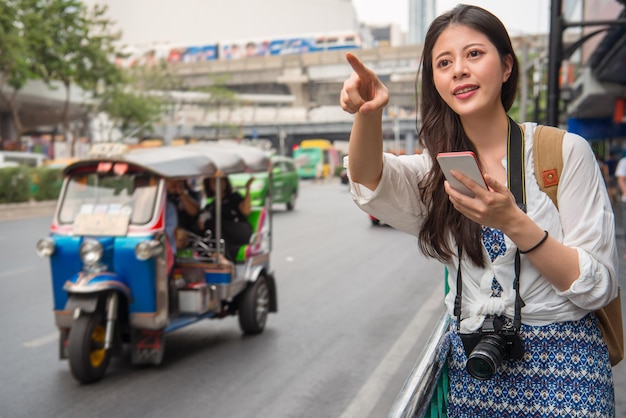 アジア人女性旅行者が通りの脇に立って前を指さし、携帯電話でタクシーを探す。