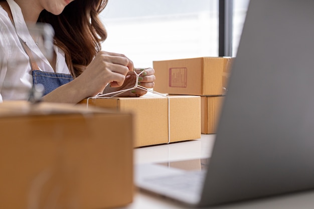 An Asian woman tying a parcel to a customer's box, she owns an online store, she packs and ships through a private transport company. Online selling and online shopping concepts.