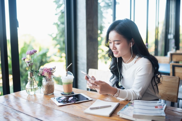 カフェでテキストメッセージを入力したり、スマートフォンで音楽を聴いたりするアジア人女性携帯電話を使用してコーヒーを飲みながらテーブルに座っているアジア人女性
