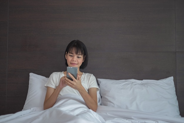 Asian woman use smartphone happily looking screen and sit in the white bed at home