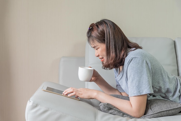 아시아 여성은 태블릿을 사용하고 소파에서 커피를 마신다