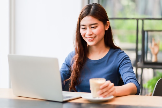 Foto la donna asiatica che per mezzo del computer portatile del computer e beve il lavoro del caffè dal concetto domestico