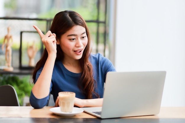 La donna asiatica che per mezzo del computer portatile del computer e beve il lavoro del caffè dal concetto domestico