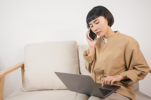 Foto donna asiatica che usa il laptop mentre si siede sul divano a casa e parla al telefono