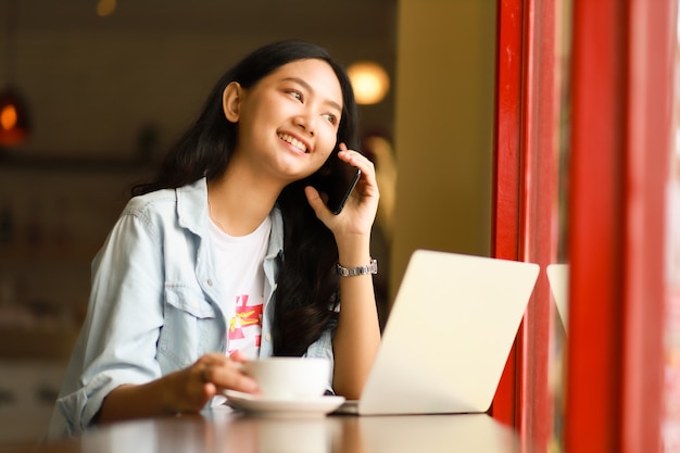 La donna asiatica che usando il computer portatile funziona e beve il caffè in caffè