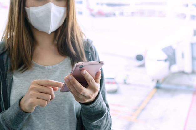 スマートフォンを使用し、空港ターミナルでサージカルマスクを着用しているアジアの女性。ヘルスケアの概念