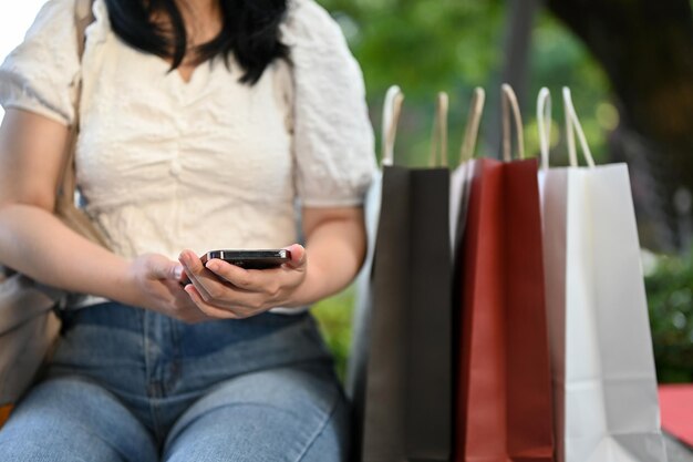 Asian woman using smartphone sitting on a bench in the shopping\
mall with her shopping bags