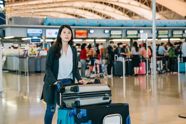 공항 카운터를 통해 걸어가는 아시아 여성은 비행기 여행을 위해 휴대 수하물로 카트를 밀고 터미널로 향합니다. 체크인을 기다리는 백그라운드에서 줄을 서 있는 사람들. 젊은 여성 노동자입니다.