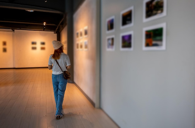 Asian woman walking through art gallery collection in front\
framed paintings pictures on white wall