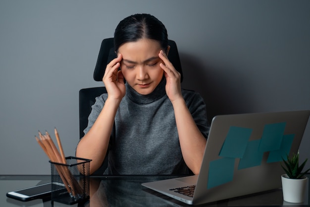 Азиатская женщина болела головной болью, касаясь ее головы, работая на ноутбуке в офисе