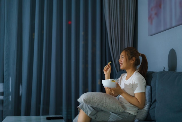 Donna asiatica che guarda film o notizie di suspense televisiva che sembra felice e divertente e mangia popcorn a tarda notte sul divano del soggiorno di casa durante il periodo di isolamento domestico.