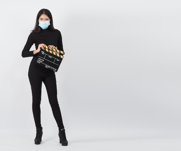 아시아 여성은 얼굴 마스크나 의료용 마스크를 쓰고 손에 검은색 클래퍼 보드를 들고 있다