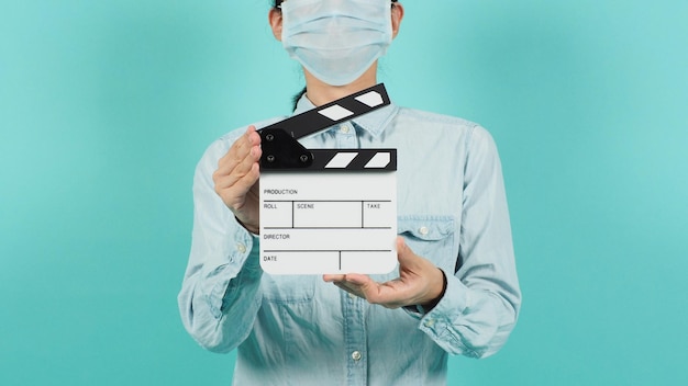 Asian woman wear face mask or medical mask and hand\'s hold\
clapper board or movie slate use on green mint or tiffany blue\
background