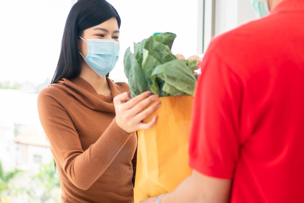 スーパーの配達人からの生鮮食品、野菜、果物のフェイスマスクとピックアップショッピングバッグを身に着けているアジアの女性。
