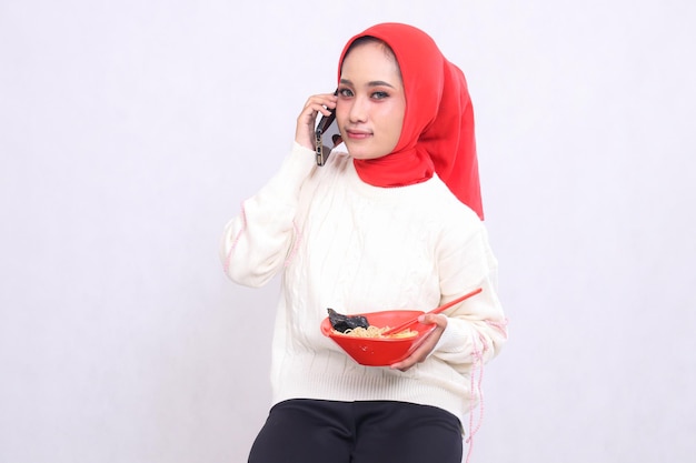 Asian woman wearing a hijab smiles on the phone with a cellphone and carries bowl containing ramen