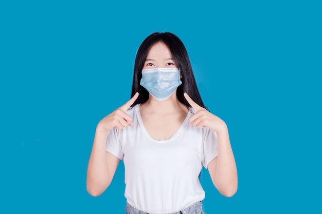 Asian woman wearing and pointing a medical face mask to protect Covid19 Coronavirus on blue background