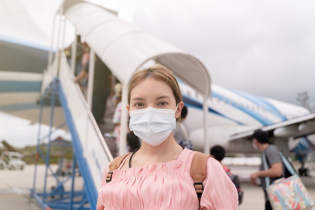 Asian woman wearing protective face mask during covid-19 virus pandemic,  walks to stair entering airplane, parking outside terminal in airport.