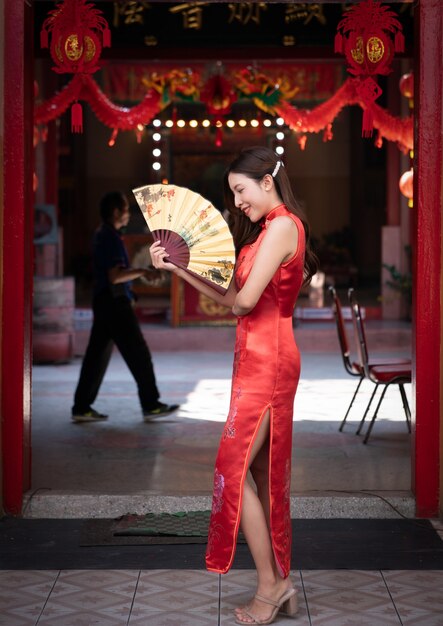 Donna asiatica che indossa un abito rosso cheongsam e che tiene un ventilatore dorato al santuario cinese. anno nuovo cinese concetto.